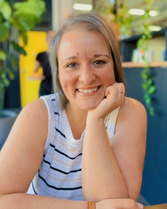 A woman with shoulder-length brown hair smiles at the camera, resting her chin on her hand, seated indoors at a table. She wears a white sleeveless striped top. Green plants and a bright yellow door are in the background.